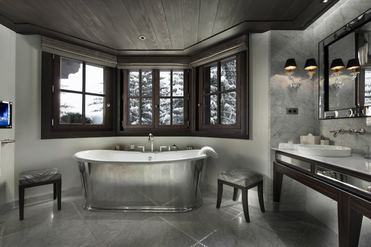 A luxurious modern bathroom with a silver freestanding tub and bay windows offering a serene view of a snow-covered winter forest.