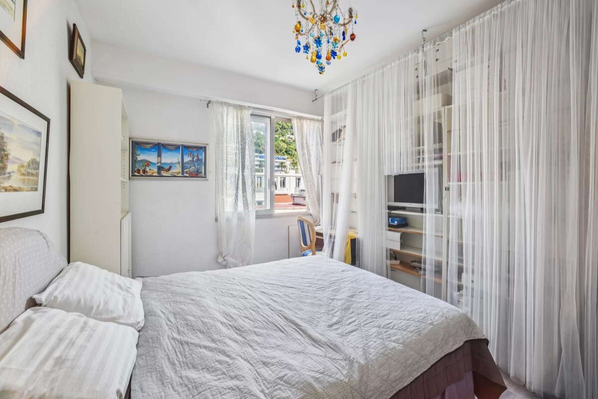 A bright bedroom features a double bed, sheer white curtains, a colorful chandelier, and a shelving unit with a TV. A window offers an outdoor view.