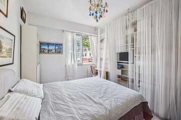 A bright bedroom features a double bed, sheer white curtains, a colorful chandelier, and a shelving unit with a TV. A window offers an outdoor view.