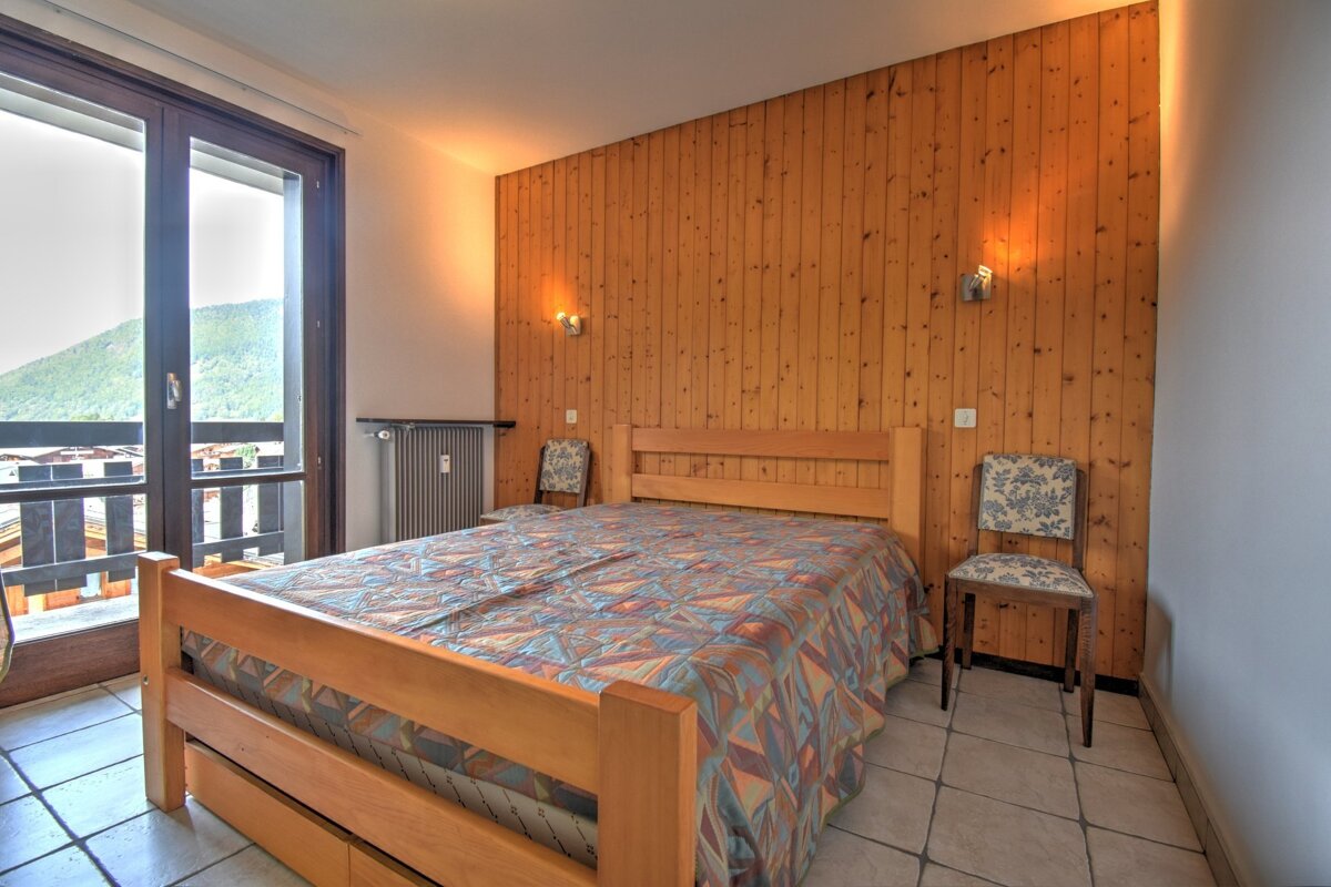 Cozy bedroom with wooden paneling, a patterned bed, tiled floor, and a balcony overlooking mountains.
