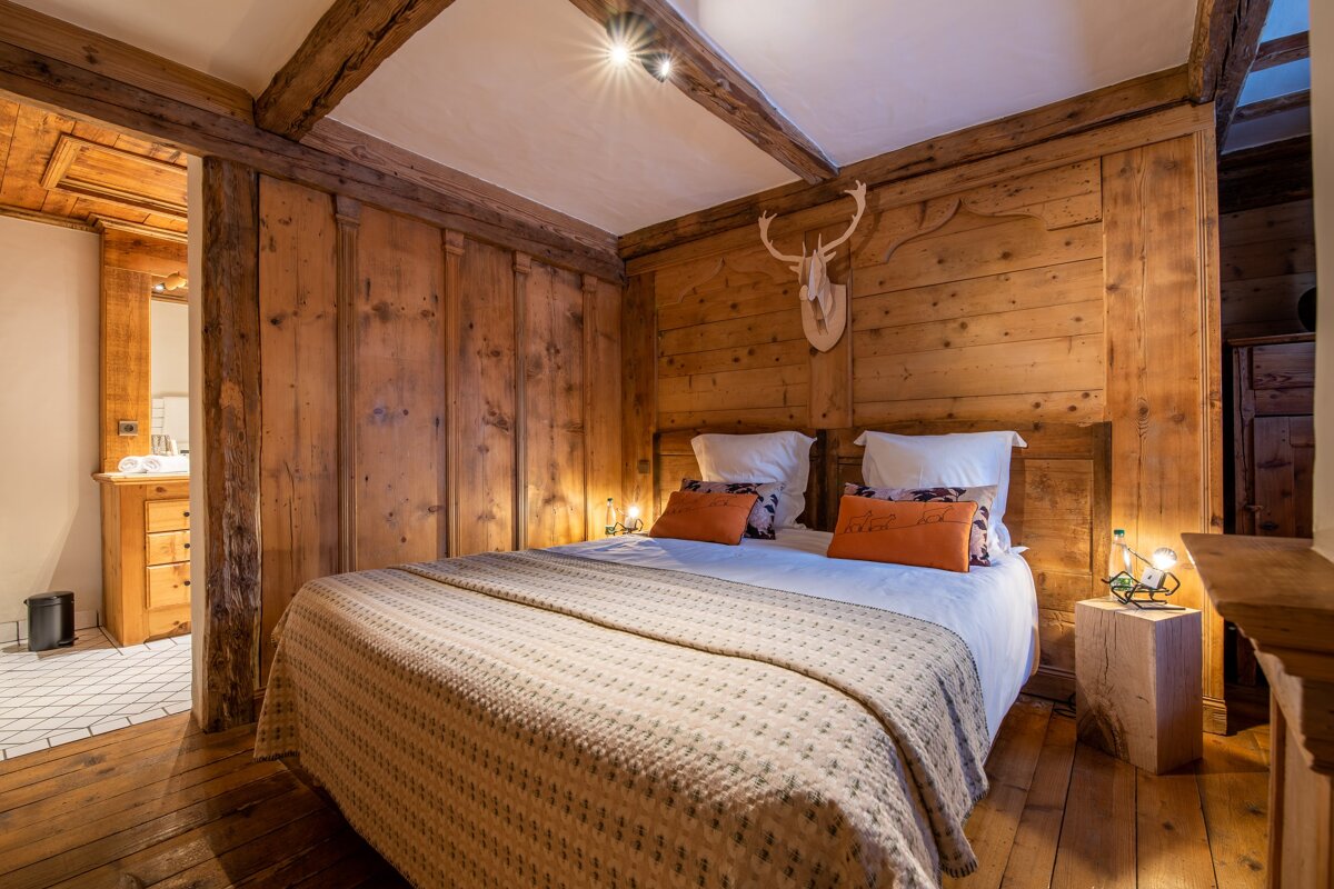 A warm, rustic bedroom with full wood paneling, exposed beams, a comfortable double bed, deer head decor, and an adjoining bathroom.