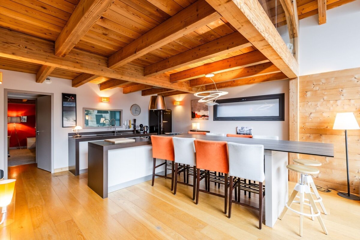 Modern chalet kitchen and dining area with exposed wood beams, a long island, and orange and white bar stools.