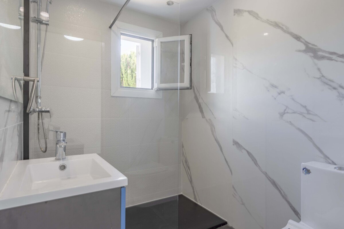 Bright, modern bathroom with white marble-look tiles, a vanity sink, shower with glass screen, and an open window for natural light.