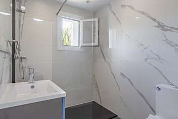 Bright, modern bathroom with white marble-look tiles, a vanity sink, shower with glass screen, and an open window for natural light.