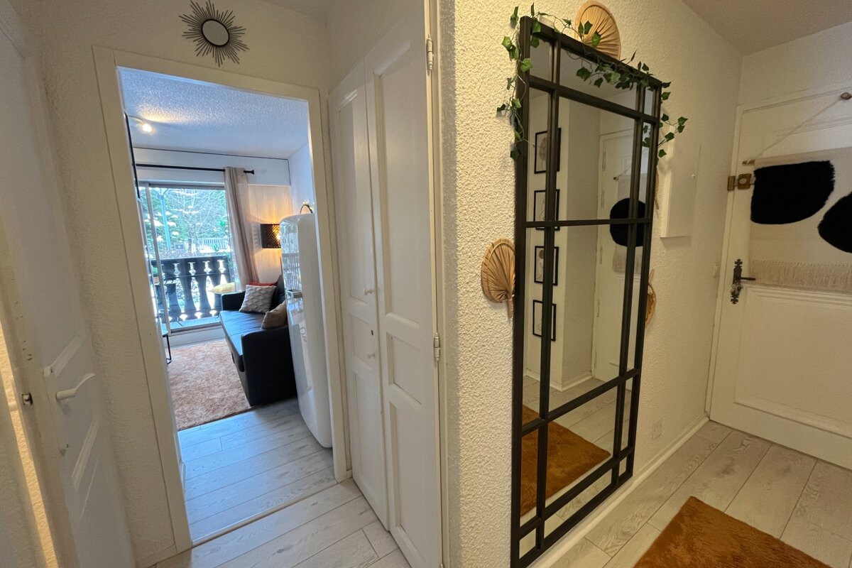 A white hallway with a large black-framed mirror adorned with ivy. Through an open doorway, a living room with a sofa and balcony is visible.