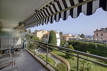 A balcony with a striped awning overlooks a lush garden, charming town buildings, and the sea in the distance under a clear sky.