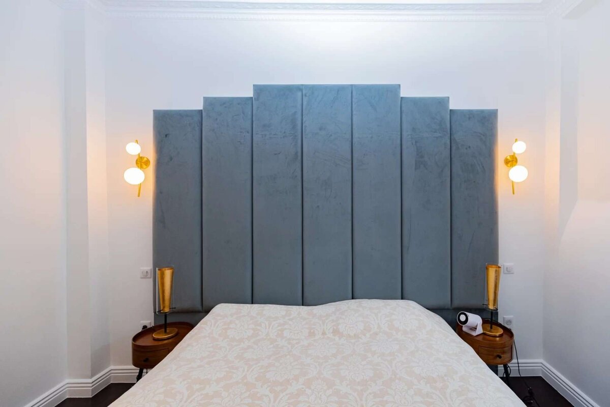 A modern bedroom features a large, tiered blue upholstered headboard, a patterned bed, and two stylish gold wall sconces flanking the bed.