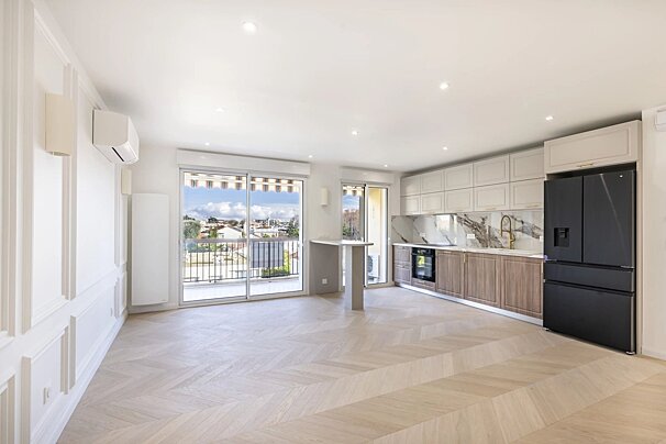Bright open-plan apartment with herringbone floors, a modern kitchen with a black fridge, and glass doors opening to a balcony view.