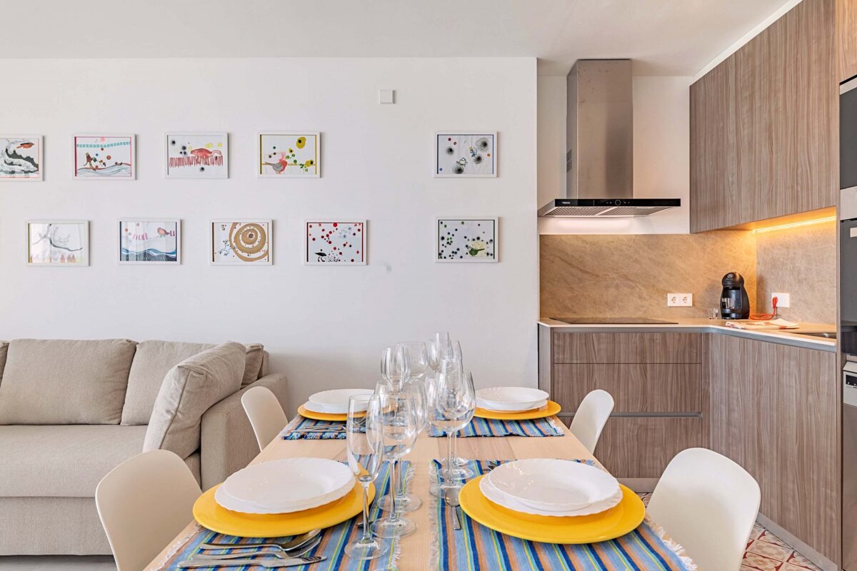 A bright, modern open-plan living space with a dining table set for guests, a sleek kitchen, a cozy sofa, and eclectic framed art on a white wall.