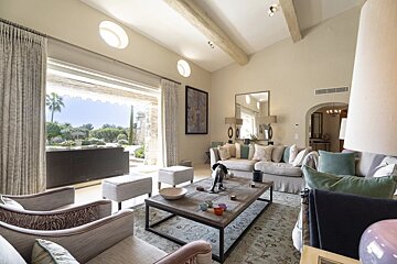 Bright, elegant living room with high ceilings, cozy neutral sofas, and large glass doors opening to a sunny outdoor patio.