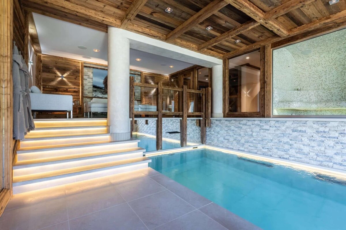 A luxurious indoor pool area with light blue water, rustic wooden ceiling, and modern illuminated steps leading to a cozy lounge, creating a spa-like ambiance.