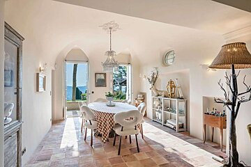 A sunny, whimsically decorated dining room with a tiled floor, overlooking a scenic view through arched doorways, centered by a crystal chandelier.
