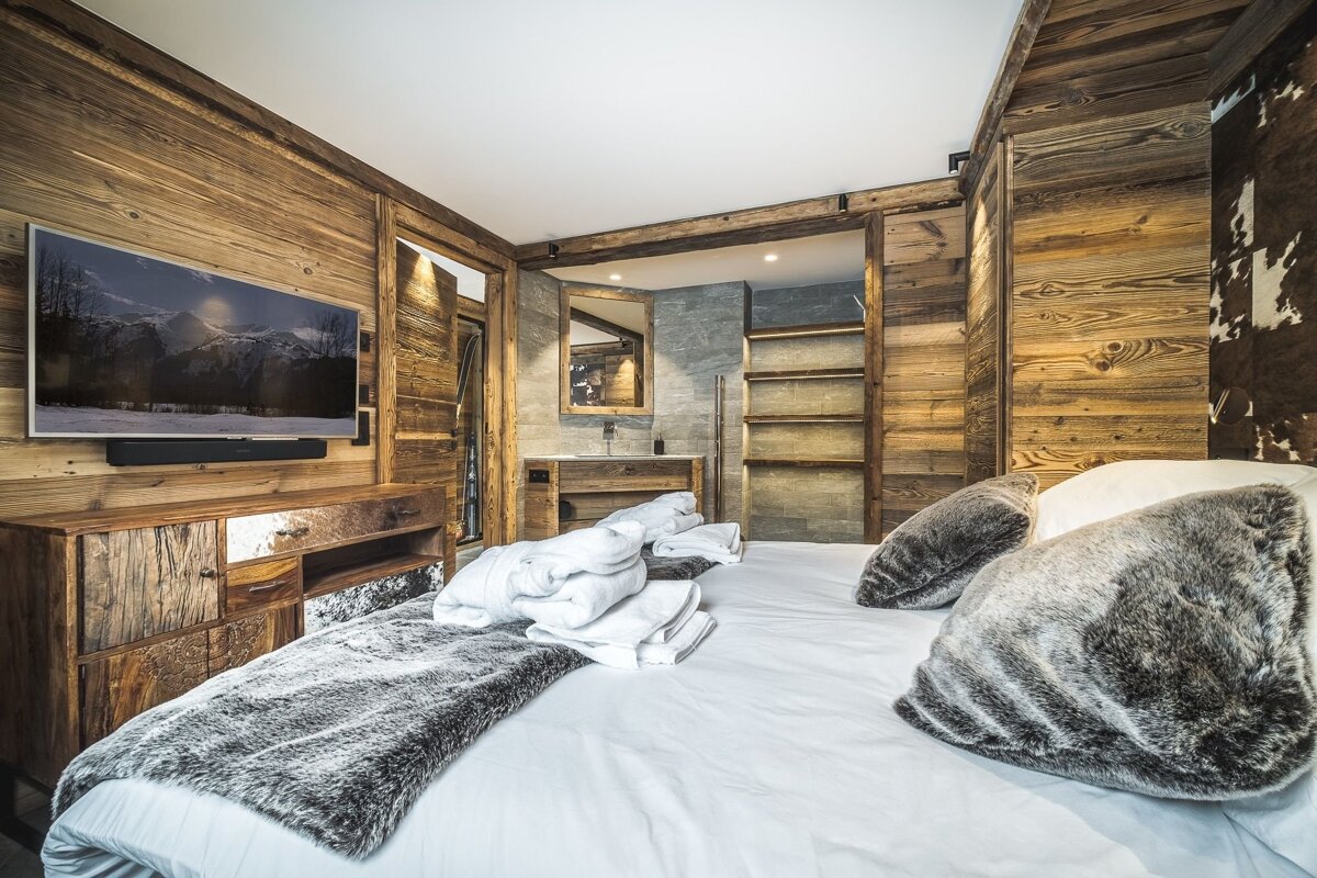 Cozy, wood-paneled chalet bedroom featuring a large bed with plush fur pillows and an en-suite bathroom in the background.