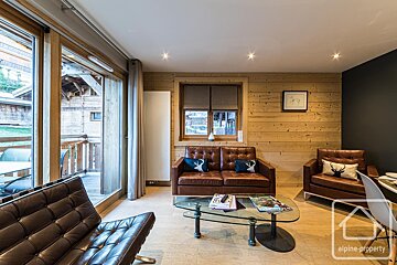 Cozy cabin living room with light wood walls, dark leather sofas, a glass coffee table, and large glass doors opening to a balcony.