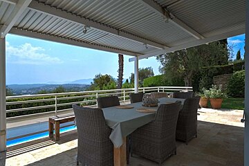 A covered outdoor dining area with a table and wicker chairs, offering a stunning view of mountains, a distant town, and a pool.