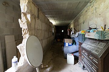 A dim, narrow, unfinished corridor with rough stone walls and a low ceiling, cluttered with old furniture and storage items.