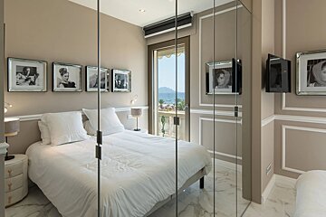 Elegant beige bedroom featuring a white bed, classic framed portraits, and glass doors overlooking a sunny sea-view balcony.