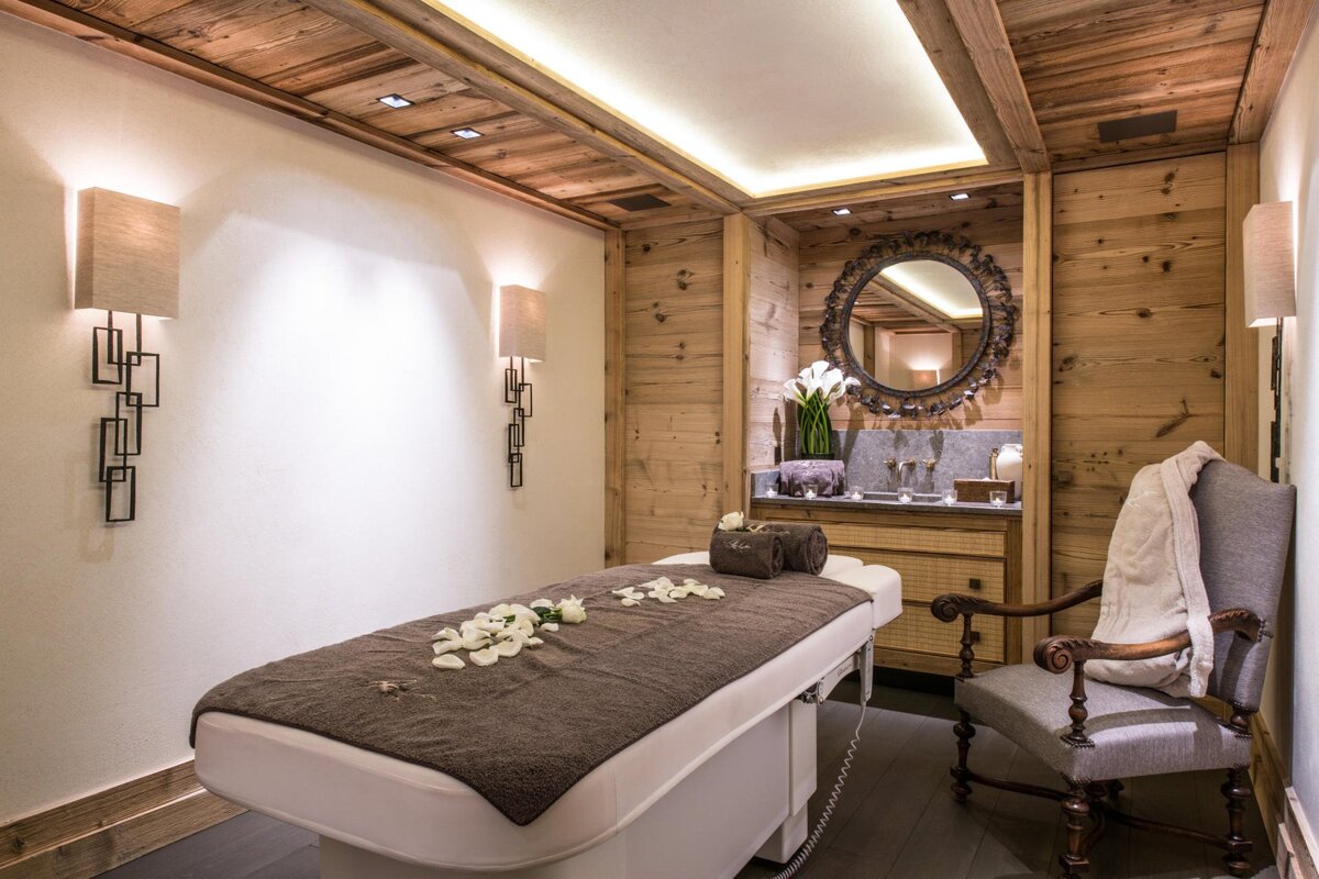 A serene spa room featuring a massage table with petals, rustic wood walls, a vanity with a round mirror, and warm lighting.