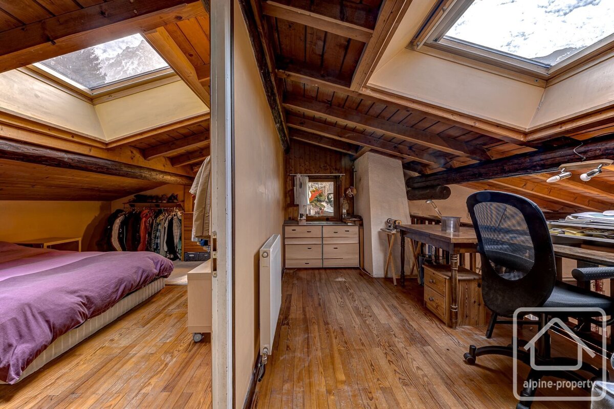 Rustic attic room with exposed wooden beams & floor. One side has a purple bed & clothes rack, the other a desk & chair. Skylights brighten both areas.