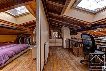 Rustic attic room with exposed wooden beams & floor. One side has a purple bed & clothes rack, the other a desk & chair. Skylights brighten both areas.