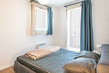 A minimalist bedroom featuring a bed with grey sheets, a pink folded blanket, and windows with blue curtains.