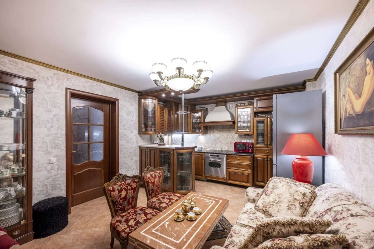 An ornate, traditional kitchen and living room combo features dark wood cabinets, floral furniture, a chandelier, and a tea set on a marble table.