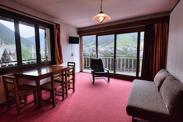 Interior of a room with red carpet, wooden table, sofa, and large windows looking out onto a mountain village.