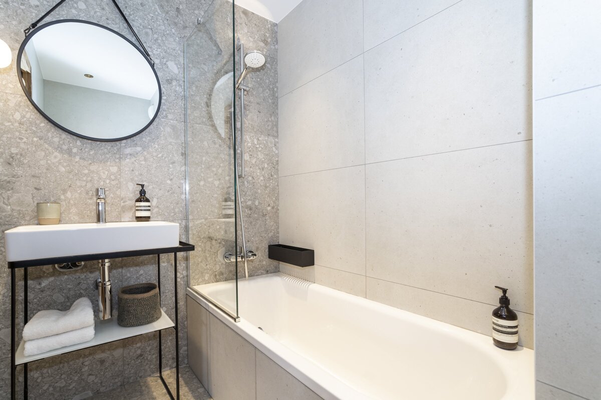 A modern bathroom features gray tiled walls, a white bathtub, a glass shower screen, and a sleek sink on a black metal stand with a round mirror.