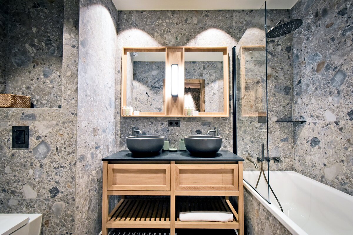 Modern bathroom with gray speckled tiles, a wooden double vanity, black vessel sinks, and a bathtub with a glass shower screen.