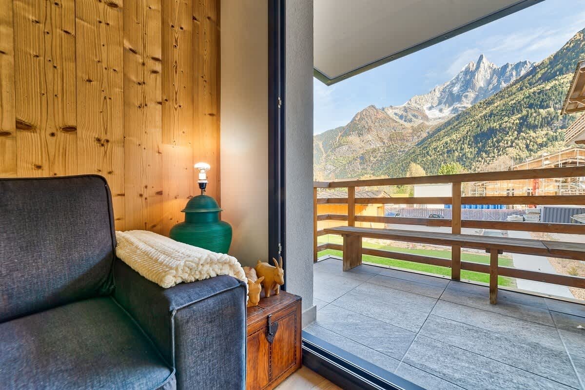 A cozy room with wood-paneled walls and an armchair opens onto a balcony with stunning mountain views.