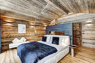 Luxurious bedroom with rustic wood paneling on walls and vaulted ceiling. A large white bed with a dark blue blanket dominates the cozy space.