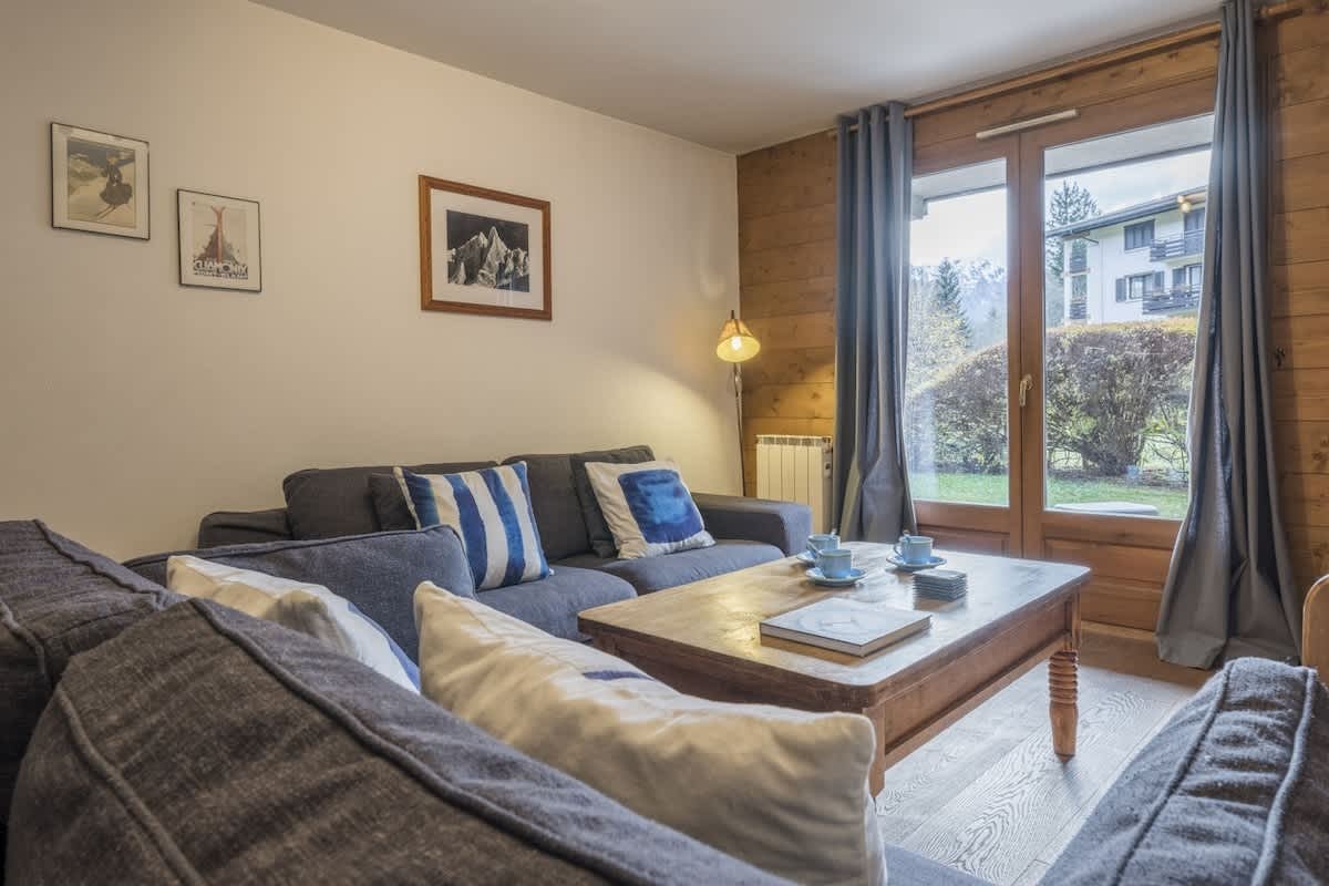 Cozy living room with grey sofas, blue accent pillows, a wooden coffee table, and glass doors looking out onto a garden.