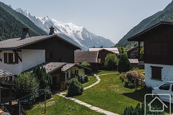 Traditional alpine village with dark-roofed houses, green lawns, and a stone path, all overlooked by majestic snow-capped mountains.