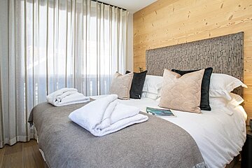 Cozy alpine bedroom featuring a wooden wall, a bed with folded white towels and pillows, and a large window with light curtains.