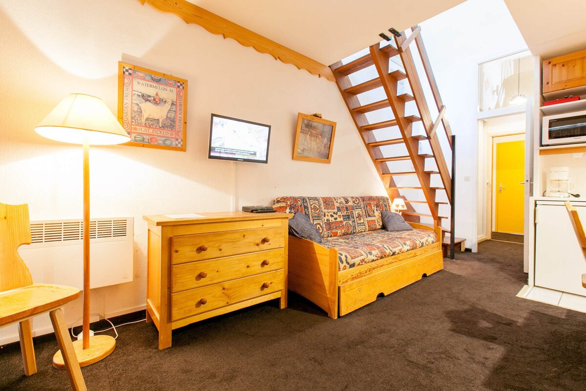 A cozy studio apartment with a patterned sofa bed, wooden dresser, wall TV, an open wooden staircase, and a compact kitchen area.