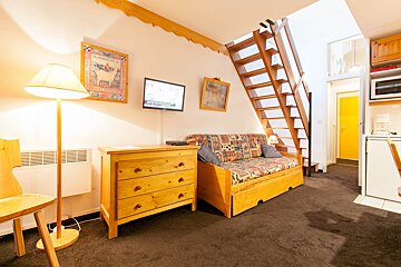 A cozy studio apartment with a patterned sofa bed, wooden dresser, wall TV, an open wooden staircase, and a compact kitchen area.