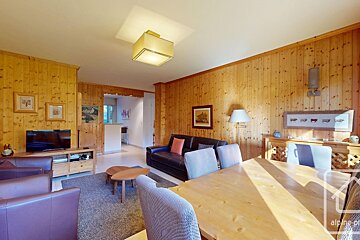 A cozy, wood-paneled living room and dining area with dark leather sofas, a TV, and a light wooden dining table and chairs. Rustic chalet style.