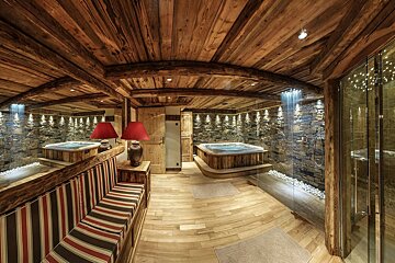 A cozy, rustic spa room featuring a hot tub, stone accent wall, wood-paneled ceiling and floor, a rainfall shower, and warm lighting.