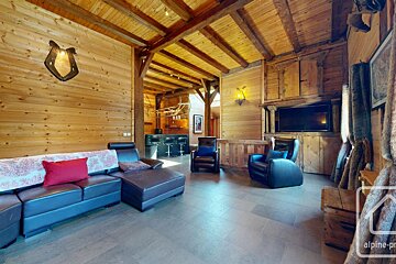 Cozy chalet living room with wood-paneled walls, beamed ceiling, dark leather sectional, armchairs, TV, and a bar area, exuding rustic charm.