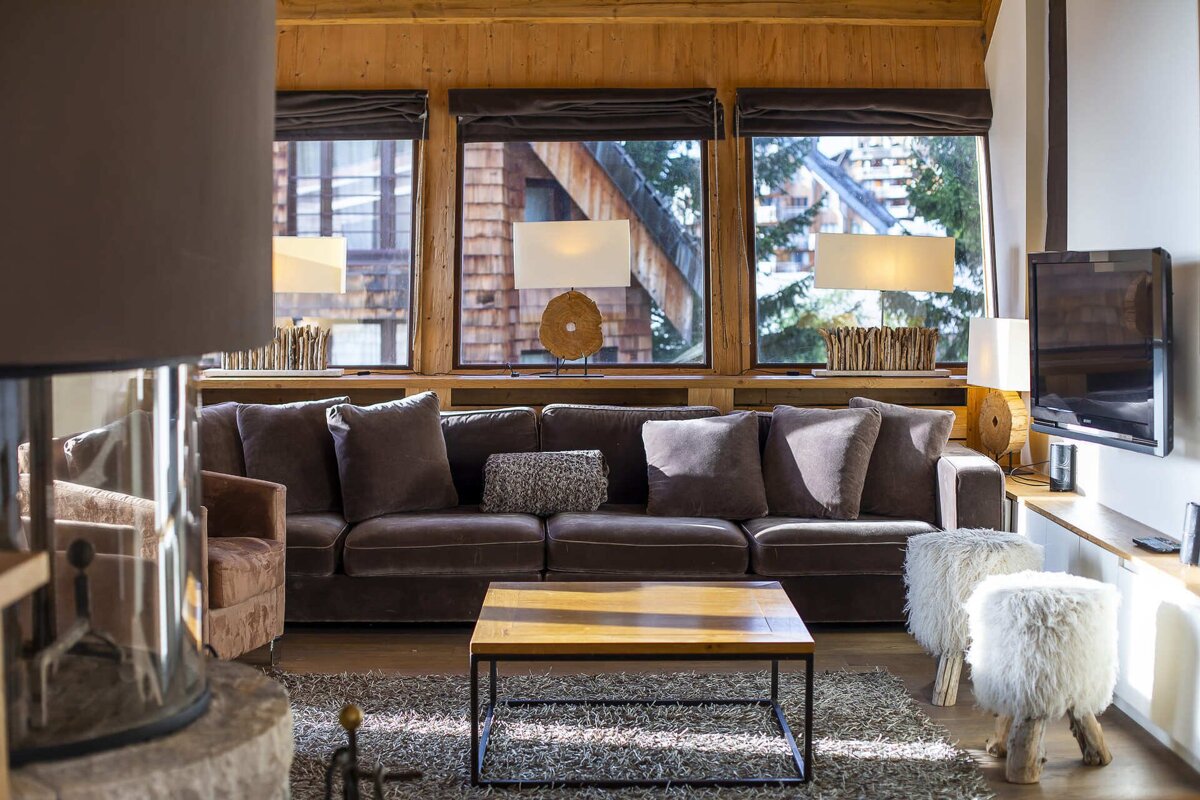 A warm, rustic living room with a large dark sofa, wood walls, and large windows revealing a winter chalet view outside.