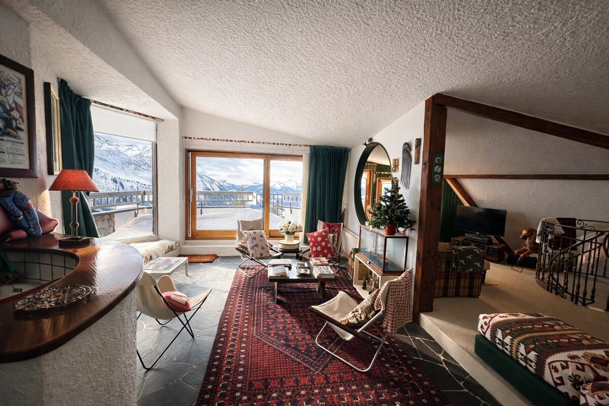 Cozy cabin living room with wooden beams, red patterned rug, butterfly chairs, and windows overlooking snowy mountains.