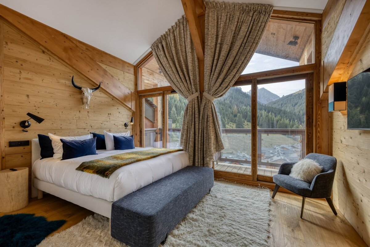 Rustic wood-paneled bedroom with a large bed, skull decor, and floor-to-ceiling windows offering a stunning mountain and forest view.