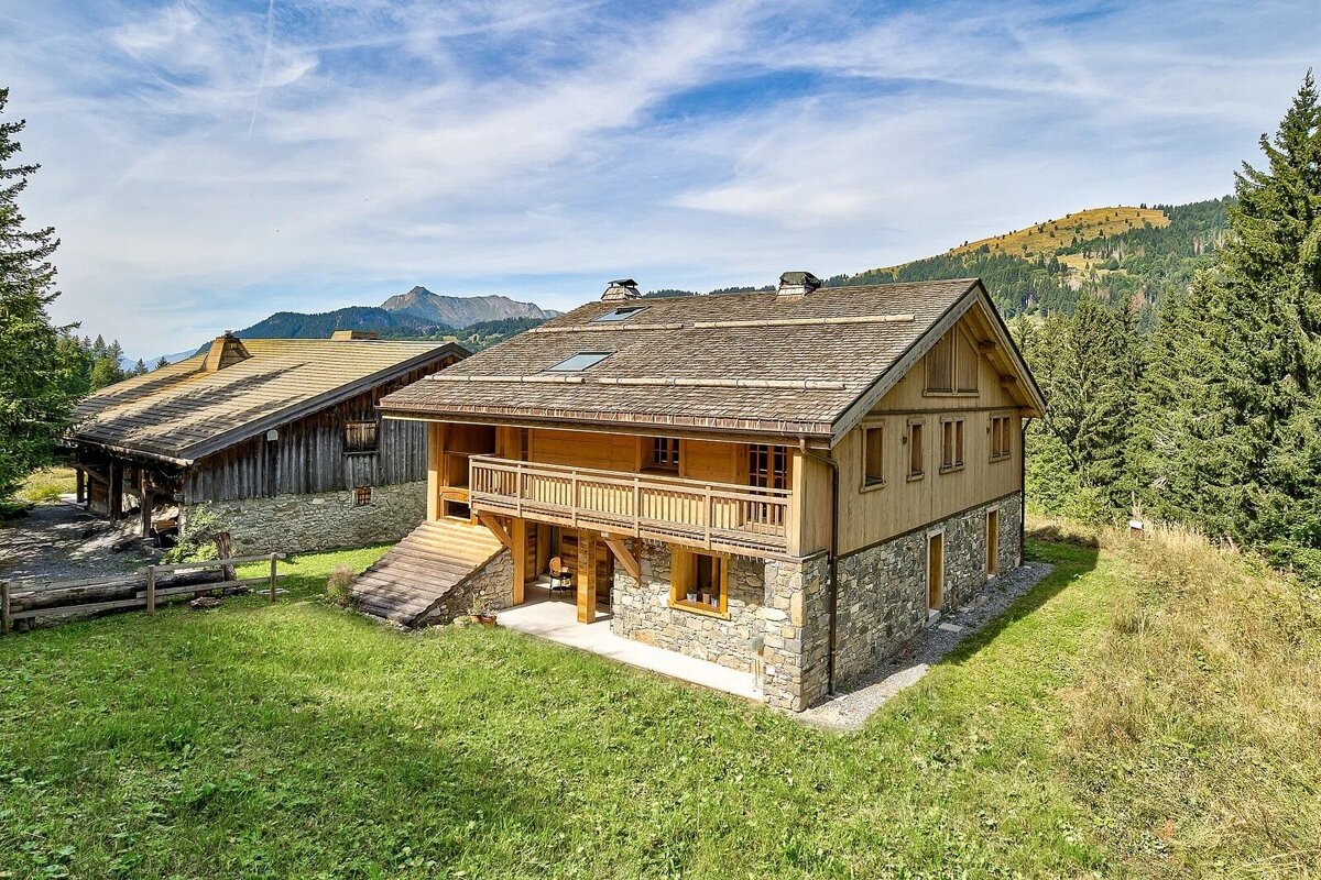 Two rustic wooden and stone chalets on a grassy alpine hillside surrounded by pine trees under a blue sky.