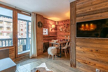 Cozy, rustic wood-paneled chalet interior with a dining area, TV, and balcony overlooking a snowy mountain town.