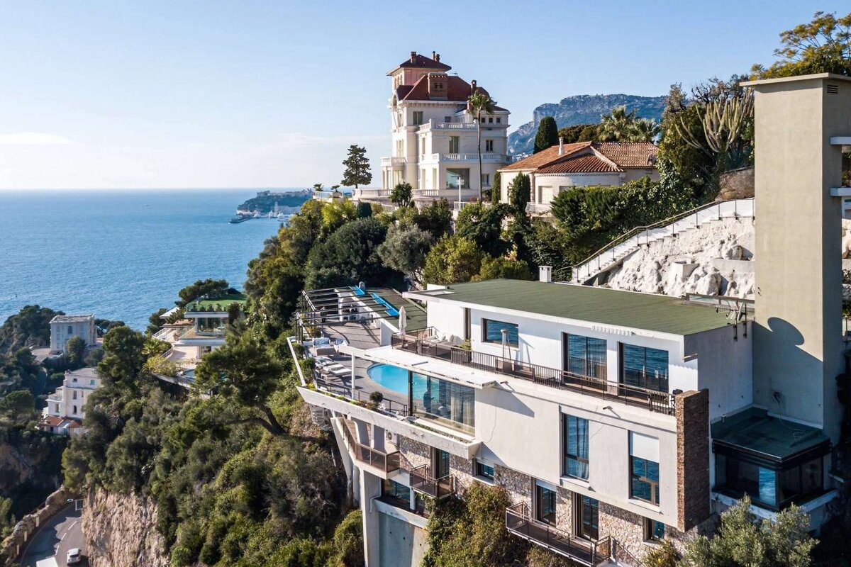 Luxurious cliffside villas with a pool terrace overlook the sparkling blue Mediterranean Sea and distant coastline under a clear sky.