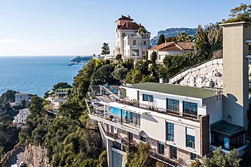 Luxury homes cling to a cliffside above the bright blue sea. Modern villas with pools blend with traditional mansions, amidst lush greenery and distant mountains.