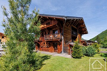A large, multi-story wooden chalet with balconies on a grassy lawn surrounded by pine trees under a clear blue sky.