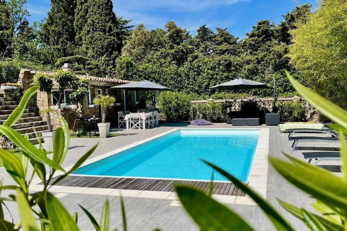 Serene outdoor pool area with dining, lounging spots, and a stone building, all amidst lush greenery under a blue sky.