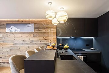 Chic kitchen blending dark modern cabinets with a rustic wood-paneled wall. Features include a breakfast bar, stylish pendant lights, and mountain artwork.