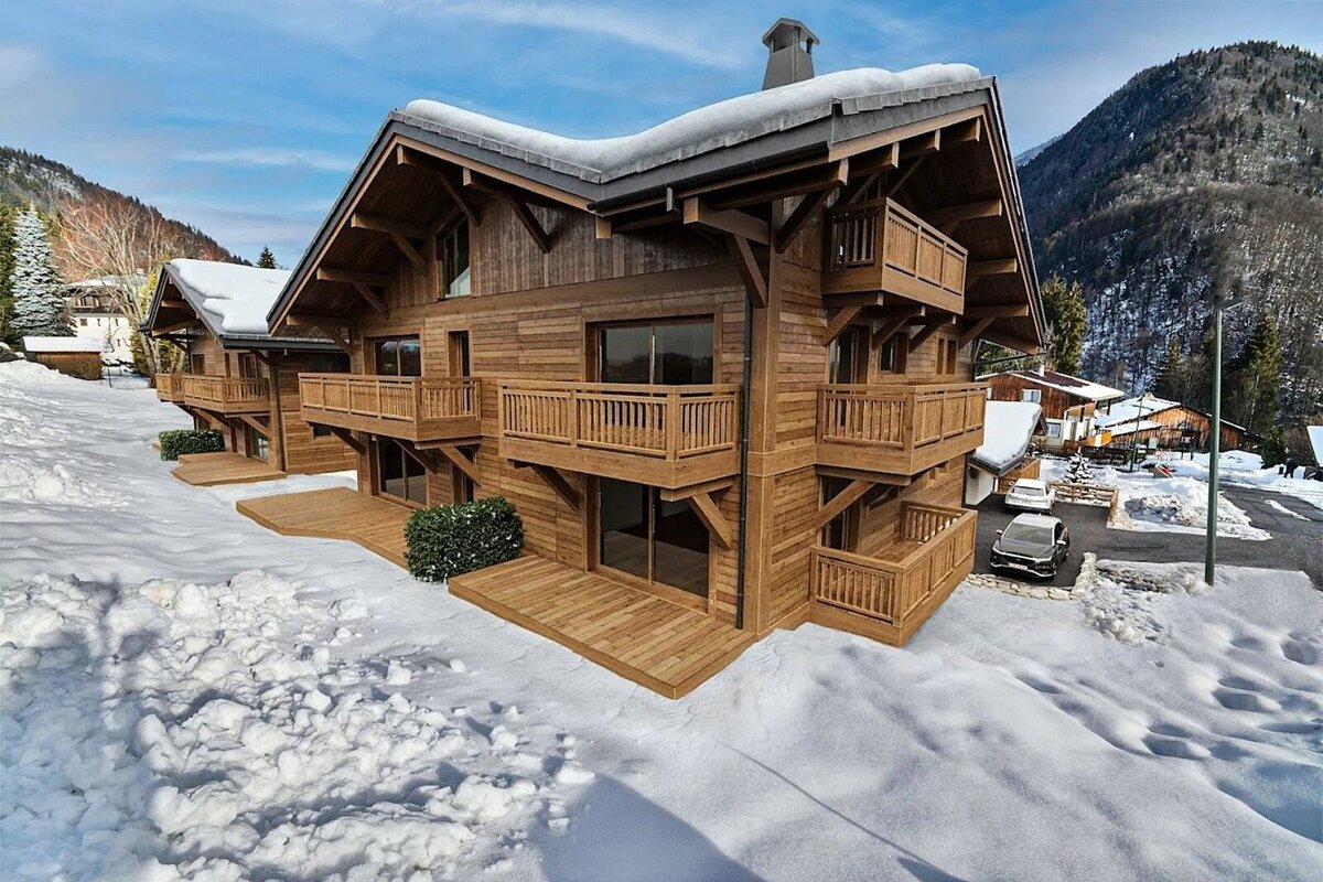 Luxury wooden chalets with balconies and snow-covered roofs in a snowy mountain landscape under a bright blue sky.